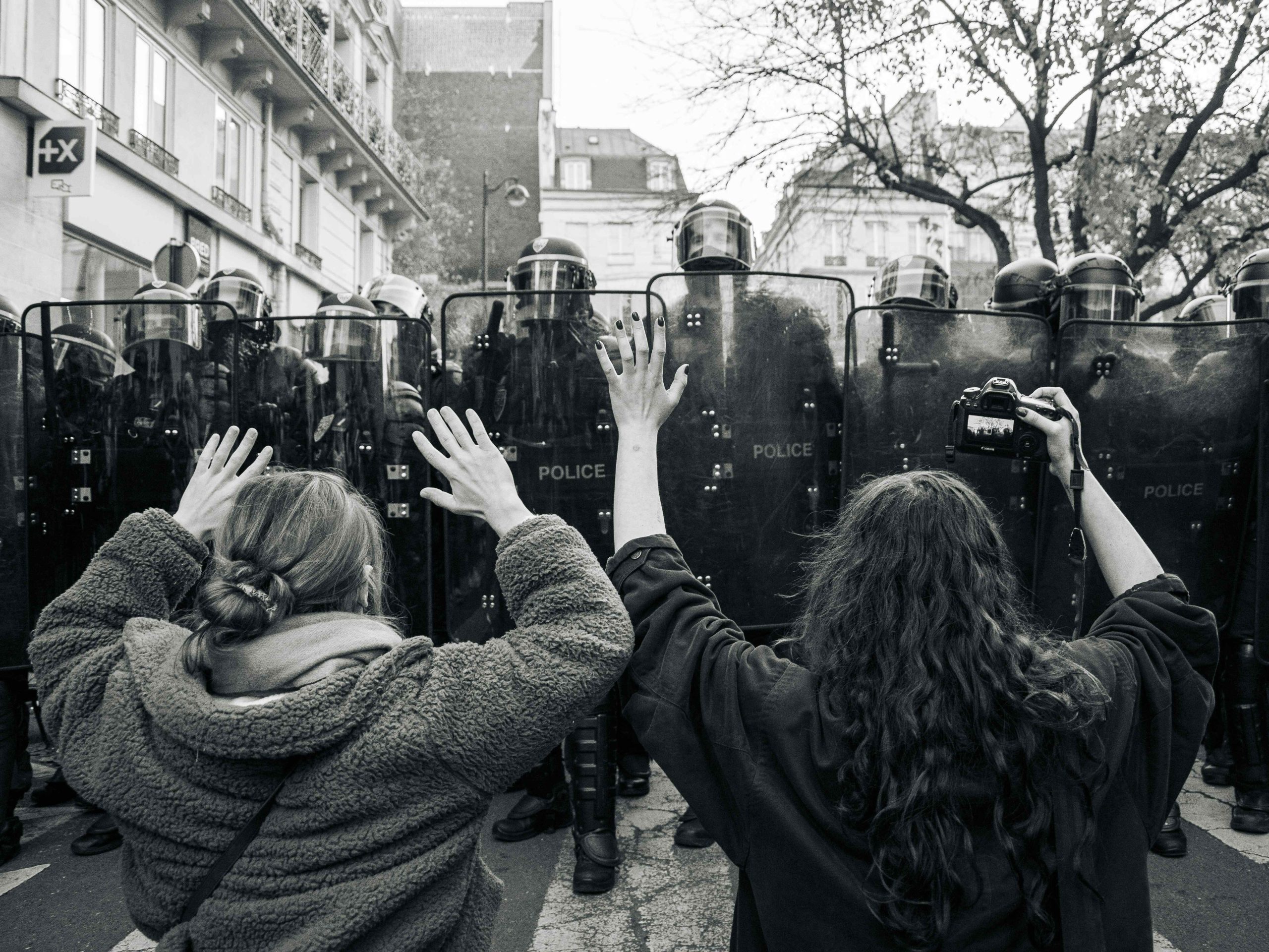 Shows two protesters with their backs to the camera lens facing a line police officers with shields.